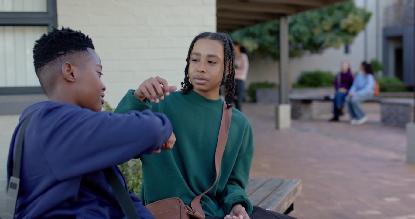 Diverse teen boys sitting on wooden bench, right boy offering object, left boy accepting as friends