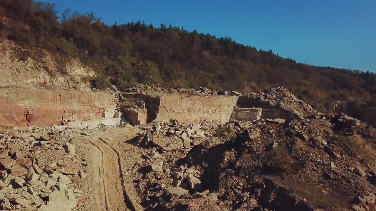 People are working around heaps of huge stones near a high and long quarry with stone rocks and with a coniferous forest on top in a warm summer day on the background of blue sky. Aerial view. Camera motion to forward.