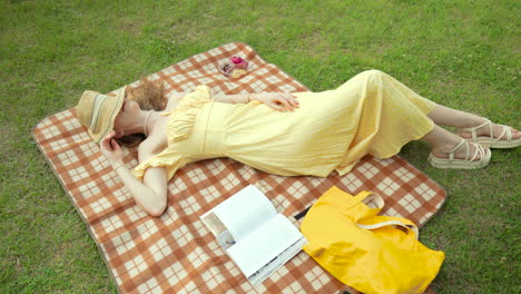 Mujer-Adulta-Caucásica-Vestida-De-Amarillo-Tomando-Una-Siesta-Acostada-Sobre-Una-Manta-De-Picnic-Con-Sombrero-De-Paja-Cubriendo-La-Cara-Relajándose-El-Día-De-Verano-En-Un-Césped-Del-Parque---Orbitando-Alrededor-Del-Movimiento