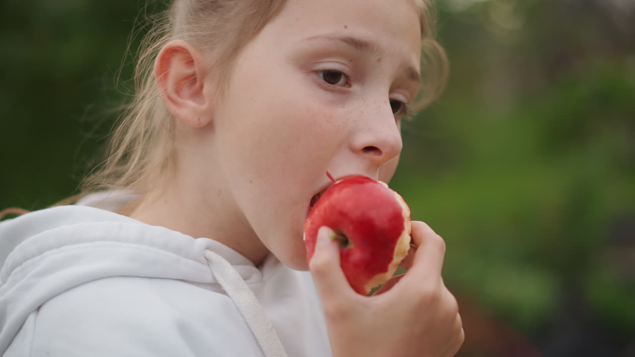 Young white girl eating apple in garden, hoodieclad schoolchild taking slow thoughtful bites of juicy red fruit, summer sunlight, candid closeup portrait conveying innocence, quiet savoring