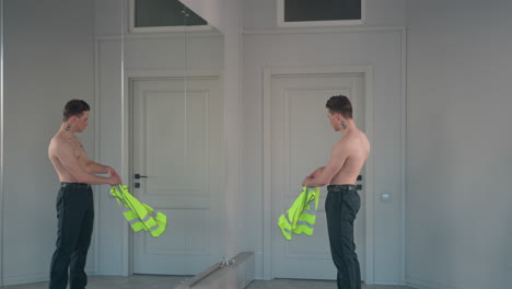 shirtless young man stands before mirror and door pulling off neon yellow jacket to reveal bold neck and chest tattoos in expressive dance pose inside bright modern studio with walls and glass