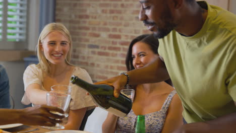 Man-Pouring-Drinks-As-Multi-Cultural-Group-Of-Friends-Enjoying-Takeaway-Meal-At-Home-Together