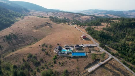 Aerial view of a picturesque hillside in Zlatibor, Sainovci, Serbia, featuring a poolside resort nestled in a lush, hilly landscape, surrounded by rolling fields, trees, and serene rural roads
