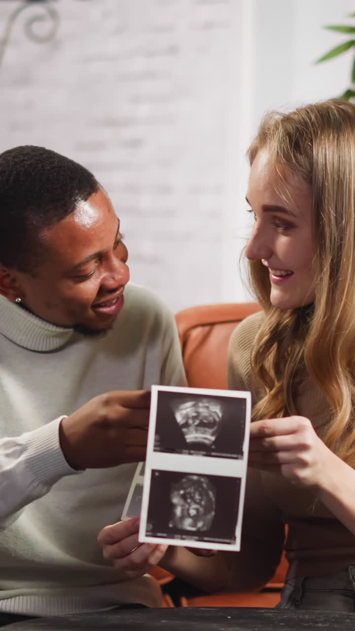 Joyful young family of black man and white lady looks at ultrasound pictures of future baby sitting on small couch in living room slow motion