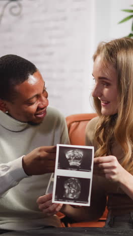 joyful young family of black man and white lady looks at ultrasound pictures of future baby sitting on small couch in living room slow motion