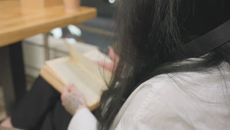 close up rear view of woman with long black hair and tattooed hand flipping page of open book while seated at wooden table with takeaway coffee cup visible in background under warm lighting