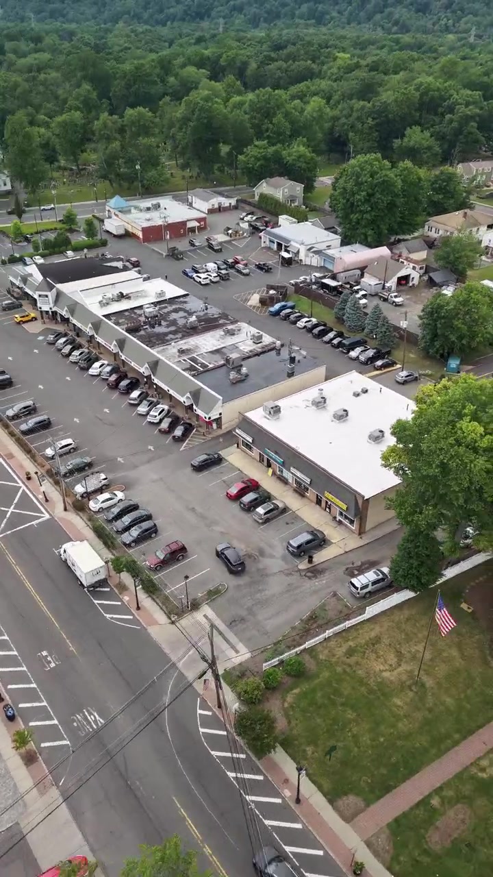 Droneoptagelse fra luften, der flyver over en typisk amerikansk småby, viser hovedgaden med kørende biler, fyldte parkeringspladser og lokale butikker omgivet af frodige grønne boligområder.