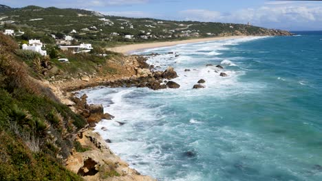 Beach-and-Phare-de-Camarinal,-Lighthouse-Camera-=-Static-shot-of-the-beach-across-2-bays-with-waves-coming-in-over-rocks-taken-from-Melia-Atlanterra-towards-the-lighthouse