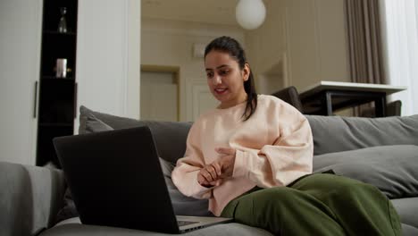 una feliz chica morena con piel oscura en una chaqueta ligera y pantalones verdes se sienta en un sofá gris y se comunica a través de video con sus colegas usando una computadora portátil durante su videoconferencia en línea en casa en un apartamento moderno y brillante