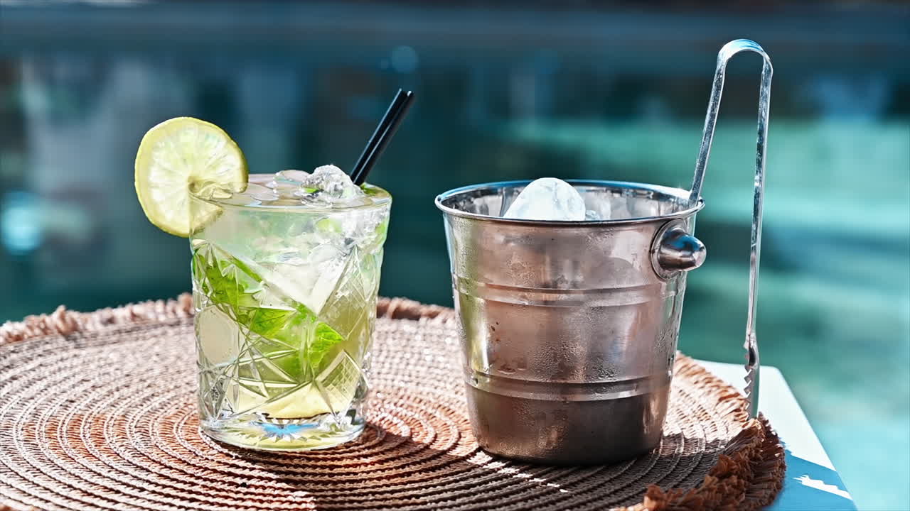 Cocktail and bucket with ice, slice of lime, ice tongs, pool on the background, Greece