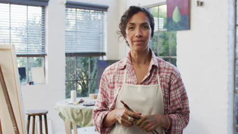 retrato de una artista biracial en delantal sosteniendo pinceles en el estudio, cámara lenta