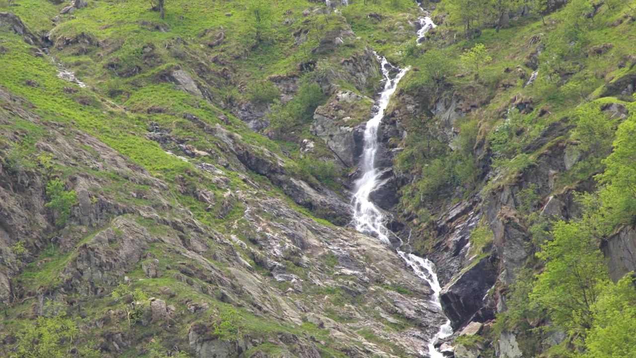 Un'inquadratura ampia statica cattura una stretta cascata che scorre lungo un ripido e verde pendio di montagna.