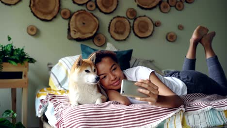 happy african american girl proud dog owner is taking selfie with cute pet lying on bed in modern apartment using smartphone. technology and social media concept.