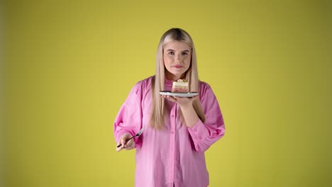 pretty blonde woman smelling a piece of delicious cake on plate with fork in the other hand, colorful studio portrait