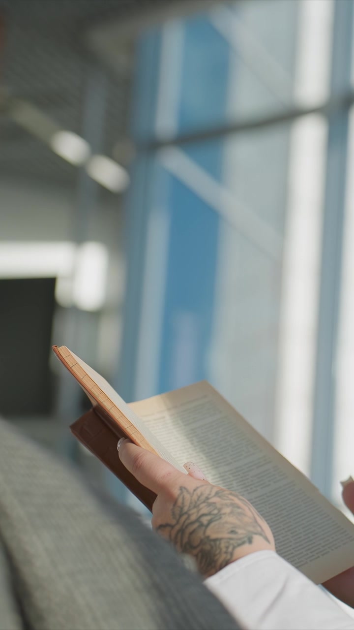 Primo piano di una mano tatuata che tiene un libro aperto su un tessuto grigio. La mano regola la presa sul quaderno, mostrando unghie di colore chiaro. Lo sfondo presenta grandi finestre e elementi strutturali sfocati.