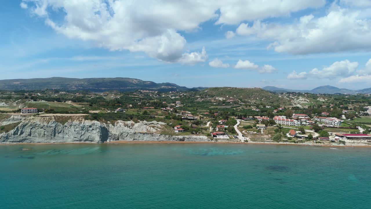 Vibrant drone capture of XI Beach with its iconic grey clay cliffs and red sandy shore, bathed in warm sunlight and overlooking the clear Ionian Sea in Greece.
