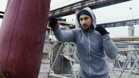 side view of caucasian bearded caucasian man in grey hoodie hitting a punching bag outdoors an abandoned factory on a cloudy morning