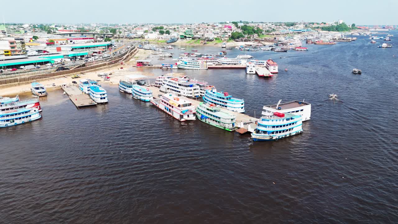 An aerial sweep over a busy river harbour in Manaus, Brazil, showing colourful riverboats and wharf activity along the Amazon River, captured in vibrant daylight