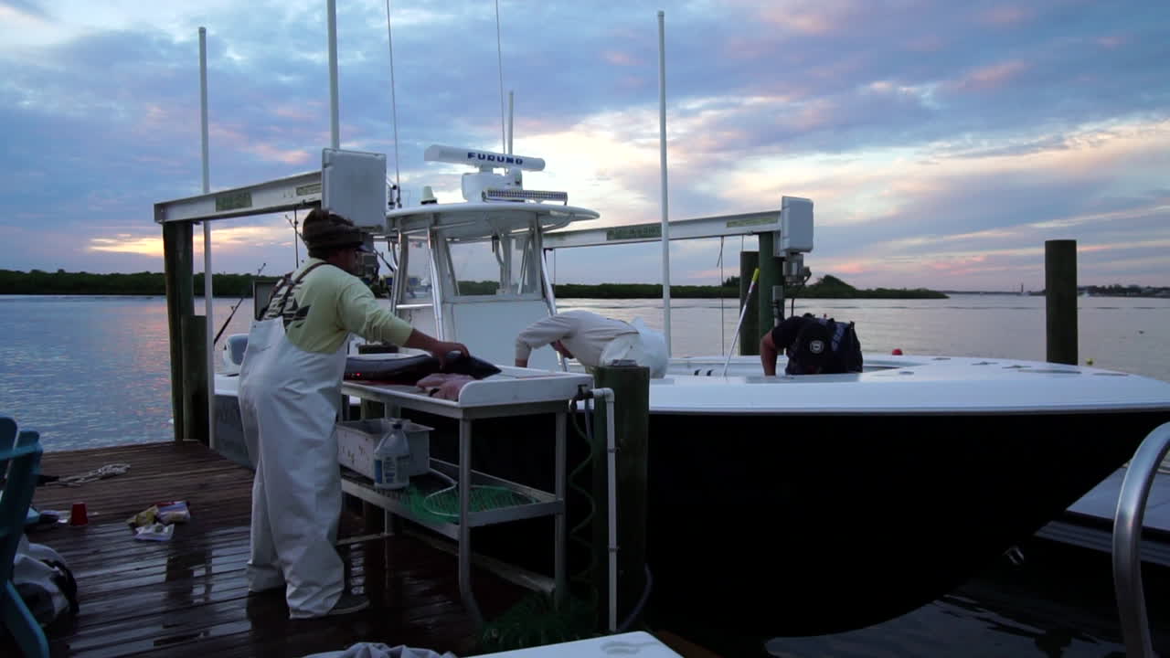 Three fishermen clean docked sport fishing boat and fillet large game fish, sunset, slow motion