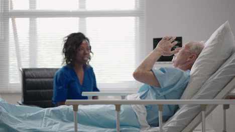 una doctora de piel oscura está hablando con un anciano paciente de sexo masculino acostado en una cama de hospital discutiendo la rehabilitación después de la recuperación.