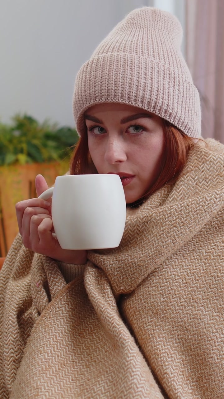 Una rossa malata indossa un cappello avvolto in un quadri si siede da sola tremante dal freddo sul divano bevendo tè caldo