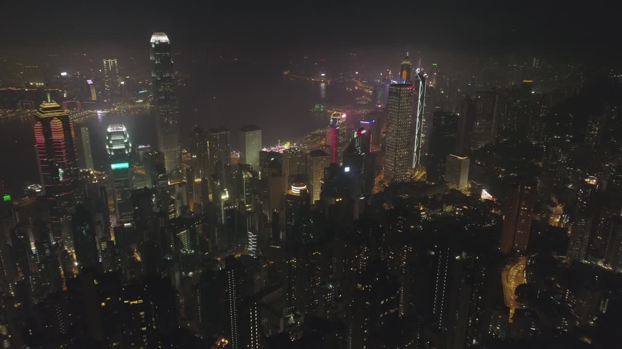 el horizonte iluminado de hong kong y el puerto de victoria por la noche. vista aérea. el dron está volando hacia adelante, la cámara está inclinándose hacia arriba. estableciendo la toma. 60 fps.