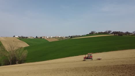 Aerial-of-red-tractor-plowing-and-preparing-agricultural-organic-soil-driving-downhill-in-farming-countryside-with-little-village-on-the-background-during-sunny-day-of-spring