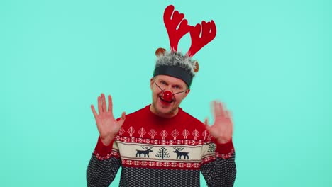 Positive-man-in-Christmas-sweater,-deer-antlers-waves-hand-palm-in-hello-gesture-welcomes-someone