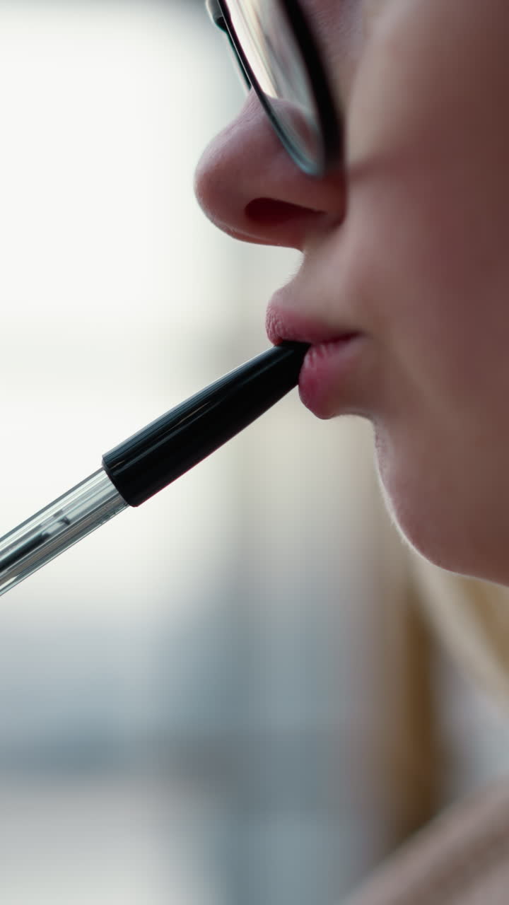 primer plano de una mujer joven girando suavemente la pluma sobre los labios, golpeando suavmente contra los labios en un gesto pensativo, con fondo borroso, capturado en un momento intelectual o creativo