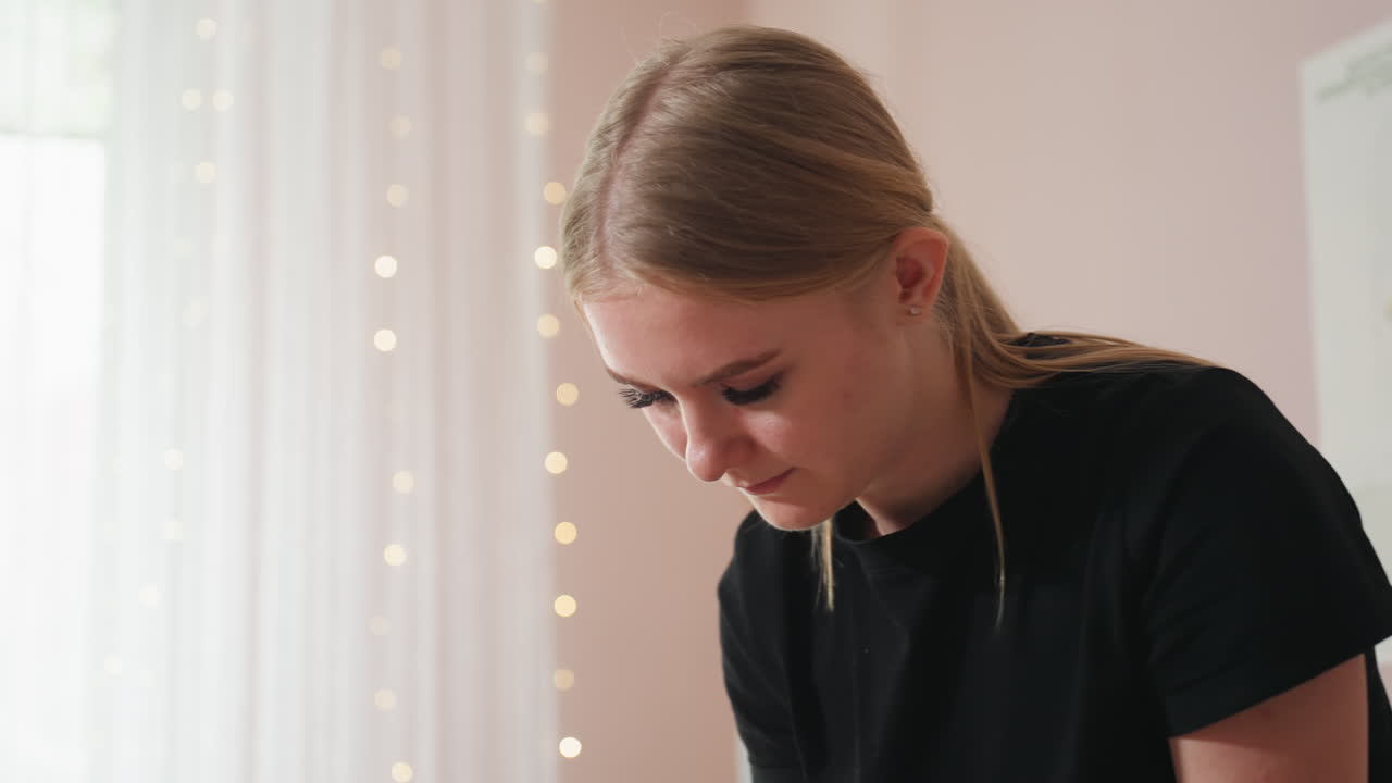 Light skin woman wearing black shirt looking down in softly lit interior, white curtain and blurry anatomical poster in background add clinical calmness and focus to the professional environment