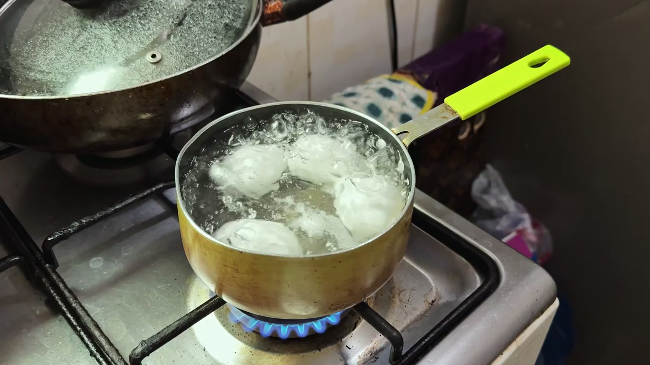 Close up of four white eggs boiling in a metallic pot on a gas stove with blue flames. Professional culinary footage for healthy breakfast preparation and home cooking lifestyle content.