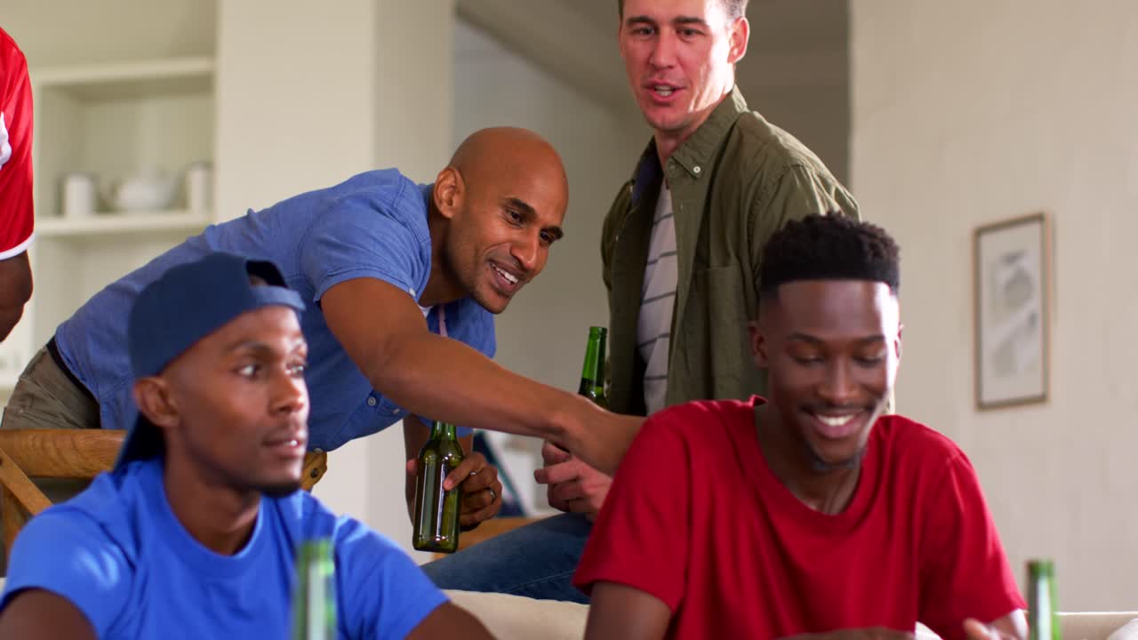 Diverse männliche Freunde zocken zu Hause, ein kahler Freund tippt einen Spieler im roten Hemd an, Gamepads und Biere.