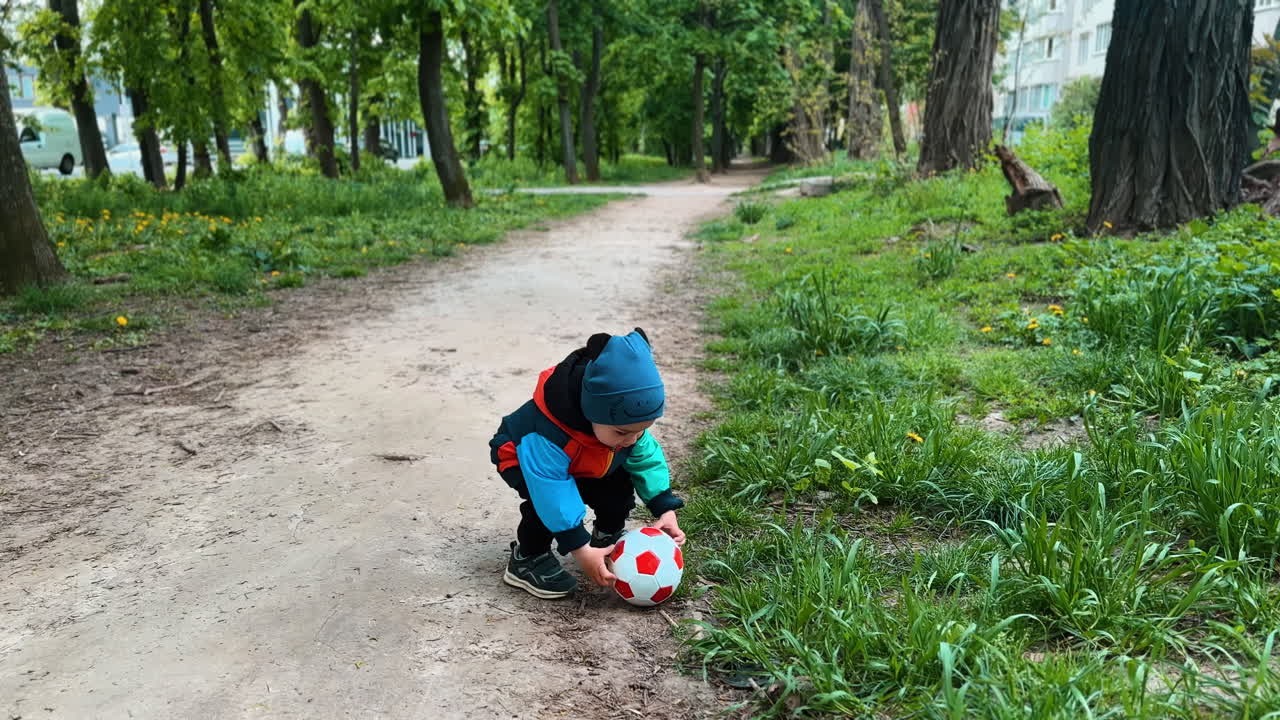 Little toddler plays with a ball in the park in spring. Baby boy in warm clothes want to lift a ball but then distracts on the stick.