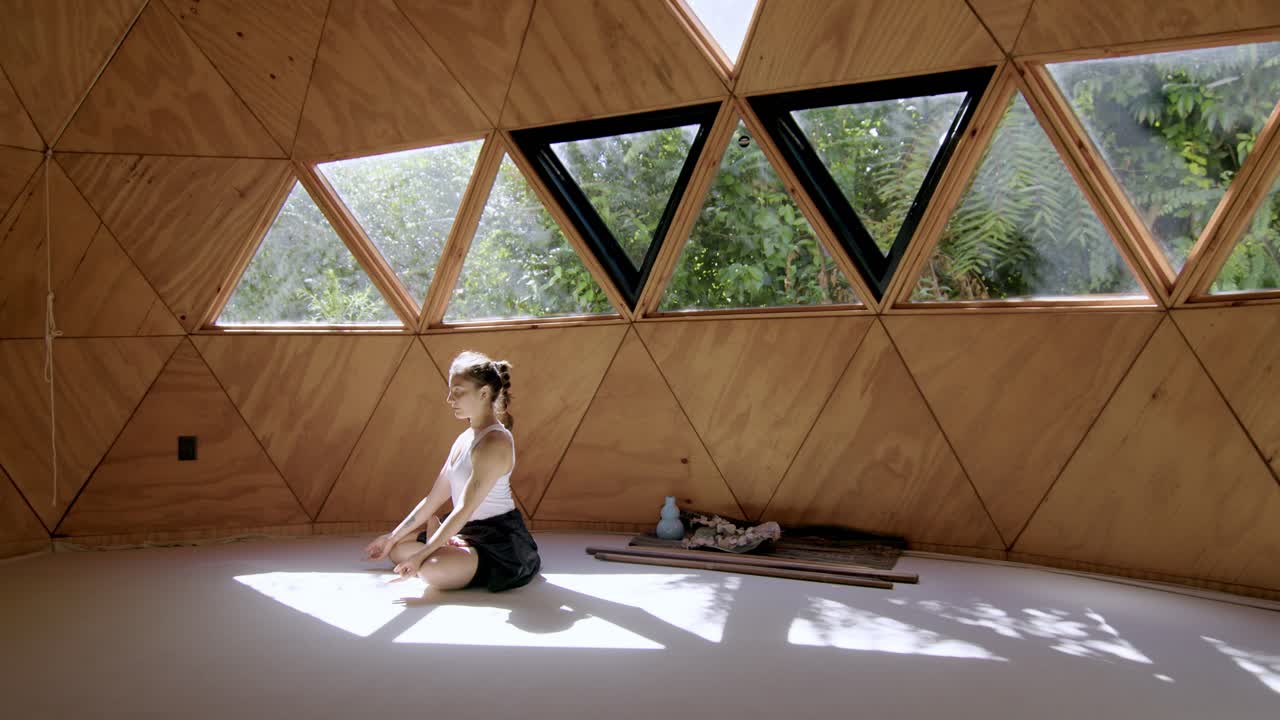 Young woman practicing yoga in a zen studio, focusing on peaceful stillness and mindfulness as tropical trees sway in windows