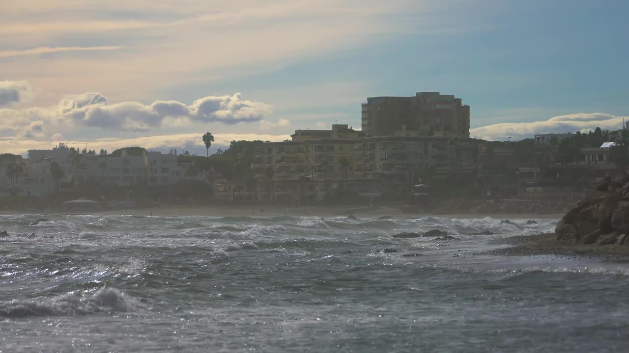 Coastal scene with waves hitting rocky shore near urban buildings under cloudy sky during daylight in southern region, showing misty atmosphere and natural movement of water and breeze