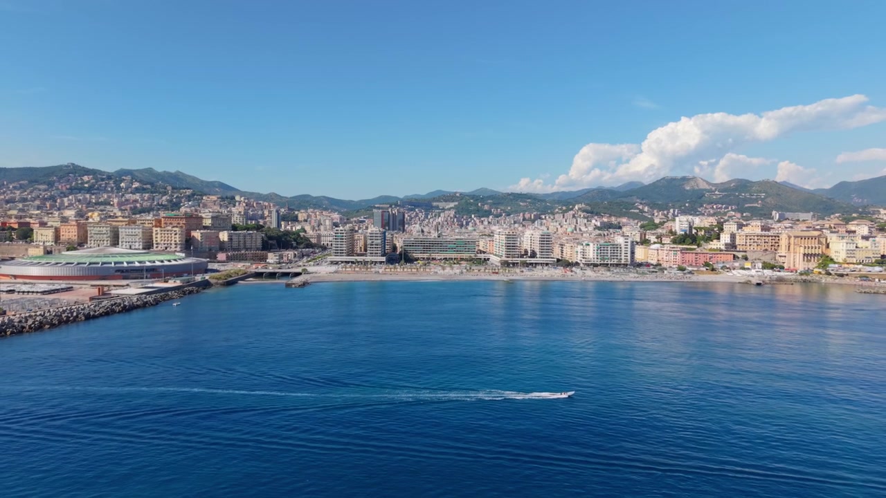 Ripresa con drone sul porto turistico di Genova con edifici cittadini, montagne e una barca sull'acqua blu.