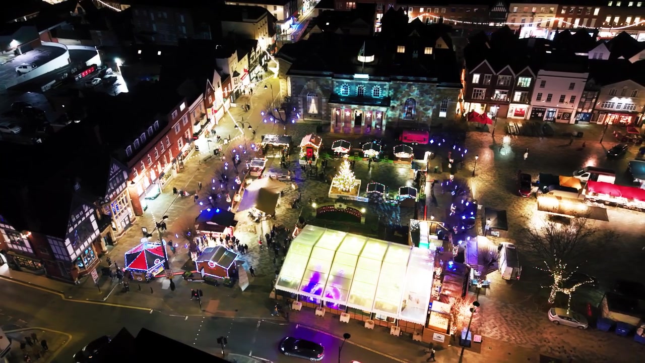 Flygfoto med drönare på natten över en vid vy av Salisburys julmarknad med Guildhallens korsvirkeshus, en julgran och upplysta stånd.