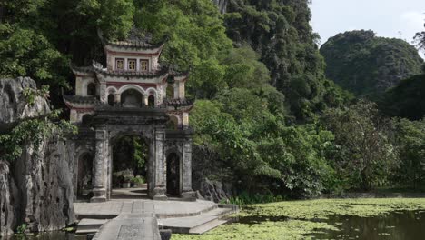 stone gate of bich dong pagoda amid lush karst cliffs and a lotus pond in ninh binh, vietnam, ornate arches and tiled roofs framed by foliage, wide static handheld shot