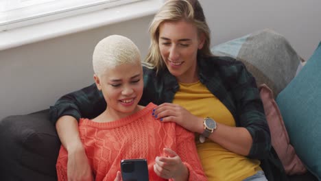 feliz pareja de mujeres diversas abrazando y usando un teléfono inteligente en la sala de estar
