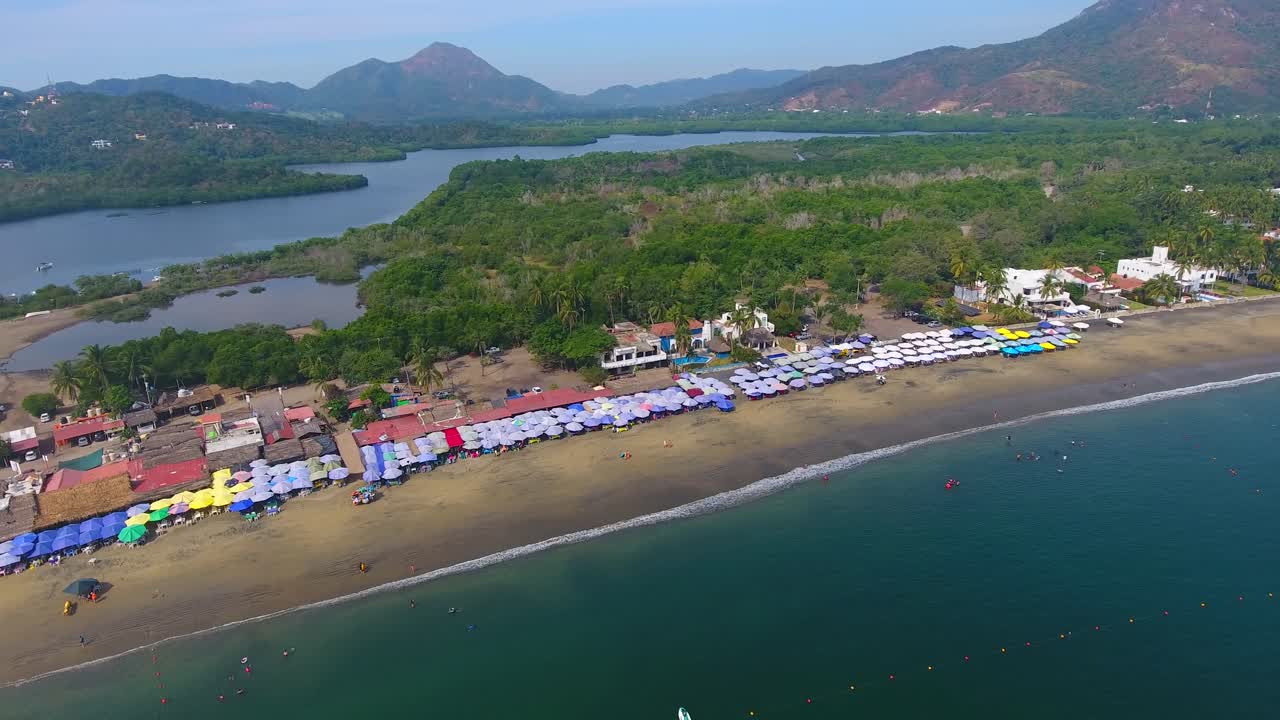 volando por la costa con vistas a una playa de arena blanca que se extiende a lo largo de una península en una isla tropical bordeada de negocios y sombrillas de playa