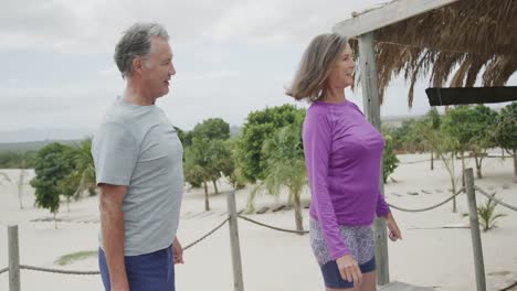Happy-senior-caucasian-couple-practicing-yoga-standing-on-beach-sun-deck,-in-slow-motion