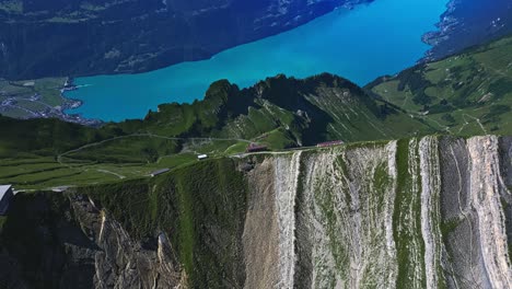 Aerial-view-of-a-hillside,-revealing-the-highest-peak-of-a-mountain,-a-pristine-blue-lake,-and-snow-capped-mountains-in-the-distance