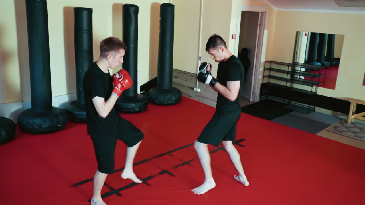 Fighters sparring on red mat inside martial arts gym, exchanging punches and defensive moves, wearing gloves and black sportswear, practicing combat skills