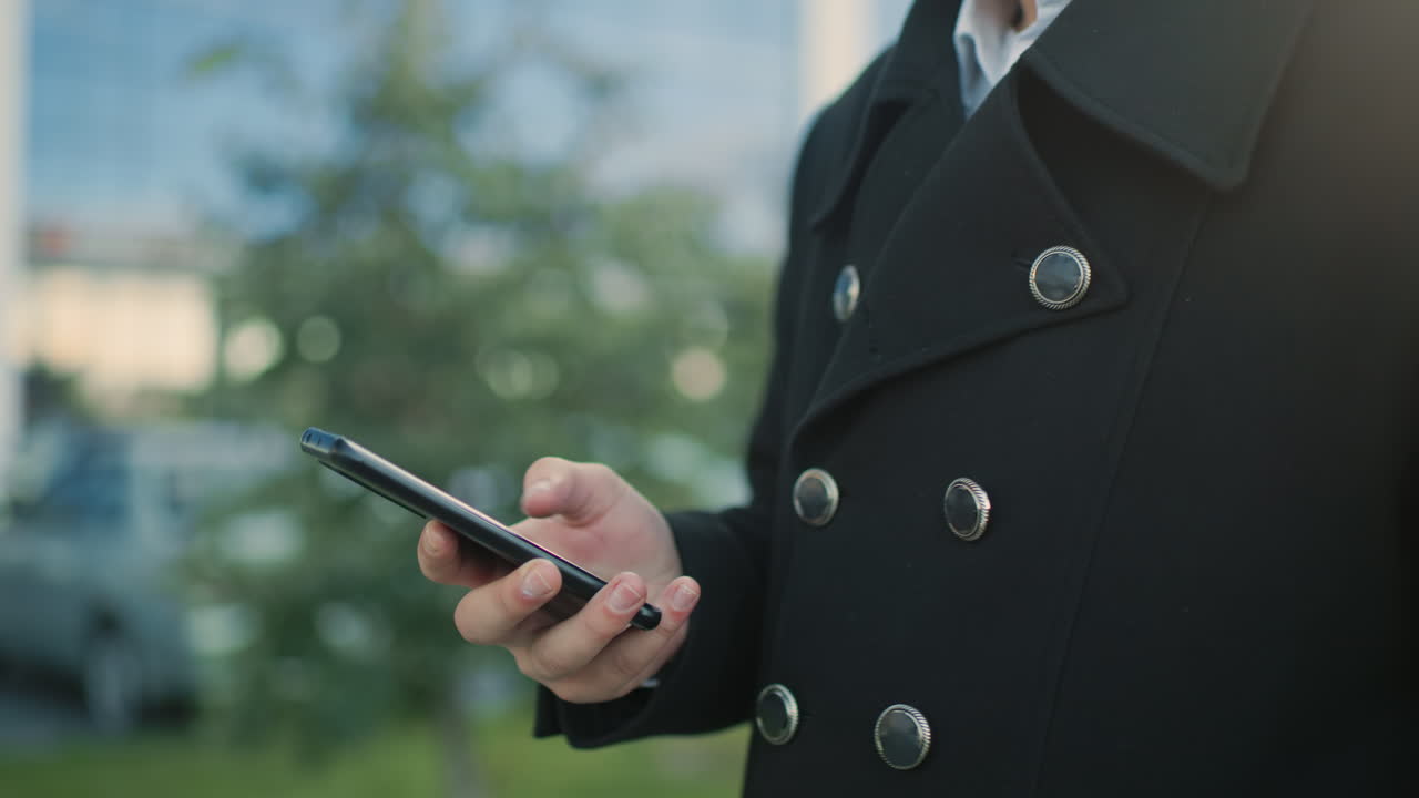 흑색 코트를 입은 개인이 야외에서 전화를 사용하는 클로즈업, 전문적인 도시 환경에서 현대적인 통신에 초점을 맞추고, 배경에는 흐릿한 초록색과 주차된 자동차가 있습니다.