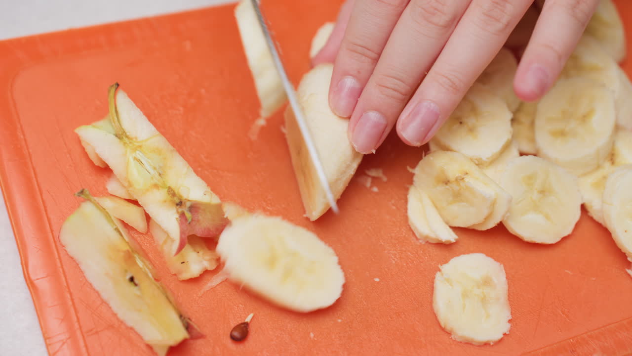 Overhead view of female hand slicing banana with knife on orange rubber board, round pieces beside apple cores, kitchen counter, clean prep vibe, fruit texture visible, shallow focus, soft light
