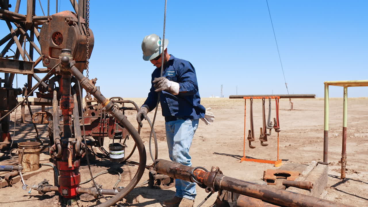 Man in helmet pulling the pipe hanging on the metal wire. Worker connecting the parts of the equipment for drilling oil or gas.