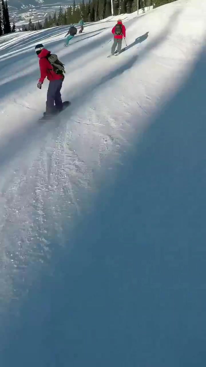 Video verticale: Inizio ripresa in discesa che insegue uno snowboarder con giacca rossa e sciatori che scendono lentamente sulla pista da sci.