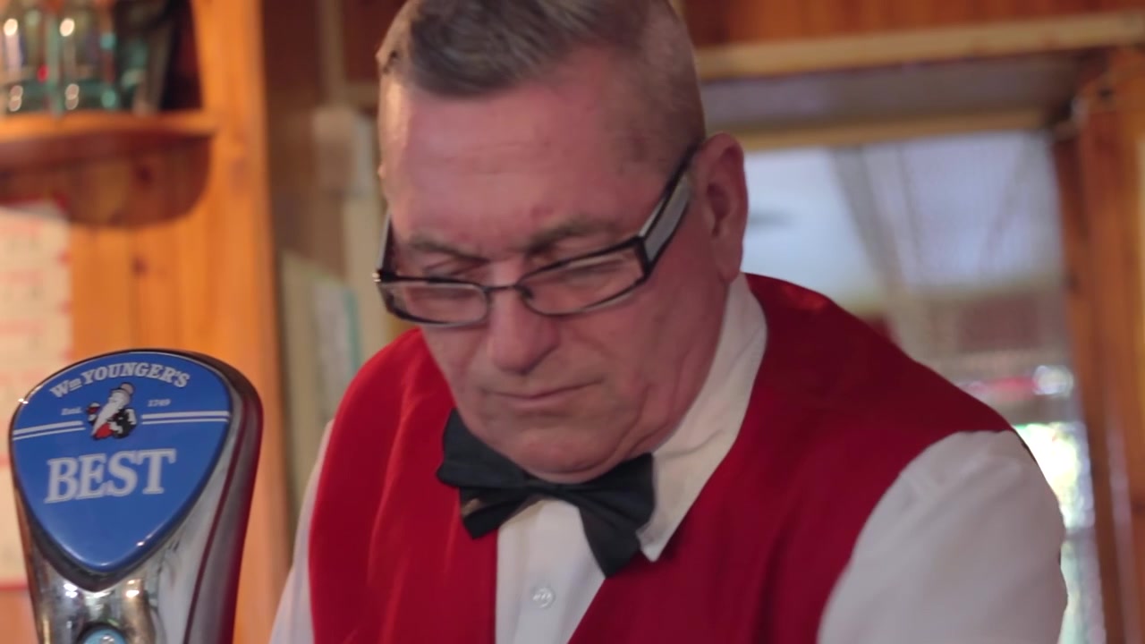 En midaldrende bartender rengør bardisken på en britisk pub. Manden bærer en rød vest og butterfly. Der er en ølhane på disken