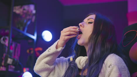 woman seated in cozy gaming chair with white headphone around neck eating popcorn as one popcorn slips from hand, illuminated by soft bulb light in a moody colorful studio environment