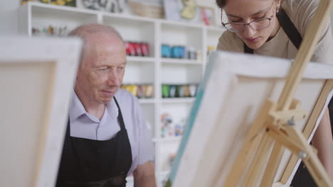 Una-Joven-Profesora-Ayuda-A-Un-Anciano-Jubilado-A-Aprender-A-Pintar-Un-Cuadro.-Hombre-Mayor-Aprendiendo-A-Dibujar